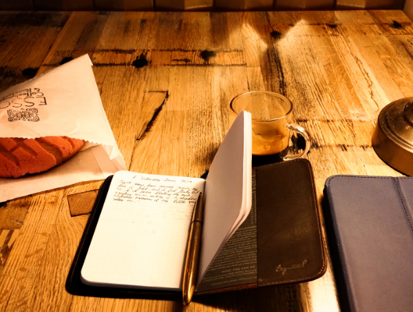 a photograph with a pastry in a paper bag, an open notebook with a brass pen, and a glass espresso cup half filled with coffee all set upon a wooden table
